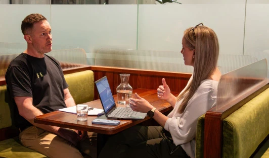 A man and a woman sitting opposite each other in a restaurant booth, talking with an open laptop in front of the woman.
