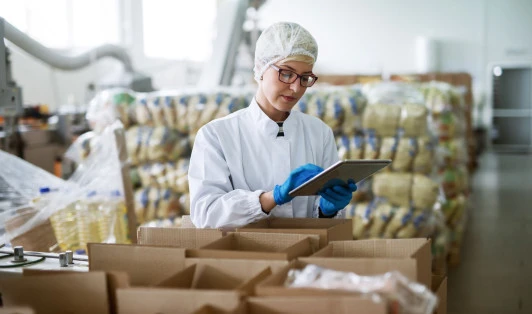 A worker in protective clothing stands in a food warehouse, looking at a tablet.