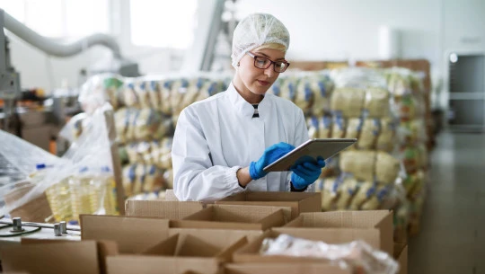A worker in protective clothing stands in a food warehouse, looking at a tablet.