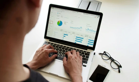Man typing on laptop looking at software, mobile phone and glasses sit beside him on a desk