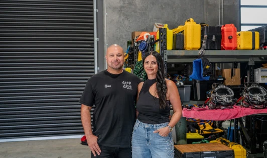 Man and woman standing next to each other in front of shelving with equipment on it inside a warehouse