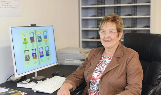 A woman sits at a desk, smiling at the camera with a computer monitor next to her.