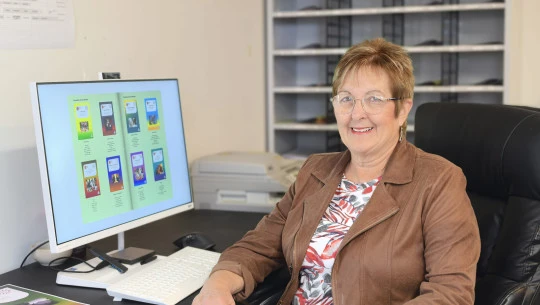 A woman sits at a desk, smiling at the camera with a computer monitor next to her.