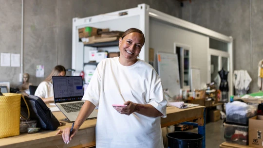 A woman stands smiling in a busy warehouse office, holding her cellphone with a laptop behind her.