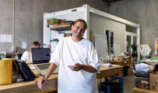 A woman stands smiling in a busy warehouse office, holding her cellphone with a laptop behind her.
