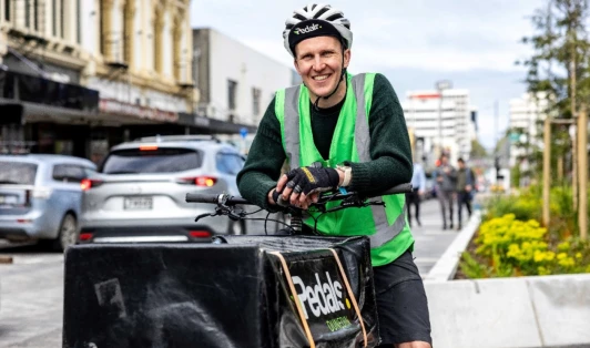 Liam Harrison stands with his cargo ebike, wearing green high vis vest, cycling gear and helmet.