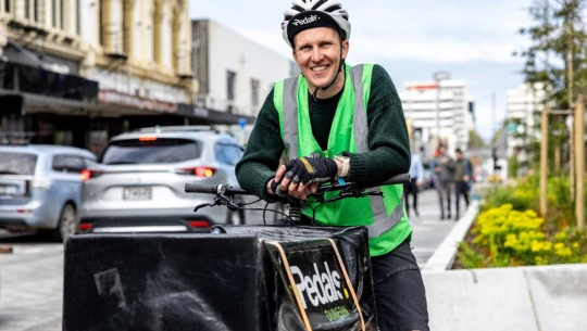 Liam Harrison stands with his cargo ebike, wearing green high vis vest, cycling gear and helmet.