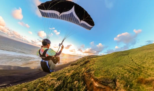 A skydiver hovers over a hillside, with a coastline to his left.