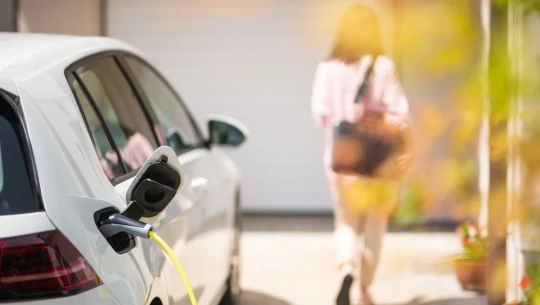 An electric car is plugged in to charge, while a woman walks away from it.