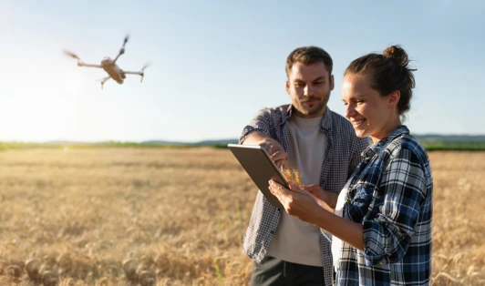 A couple stand in a field, looking at a tablet, while a drone flies above them.