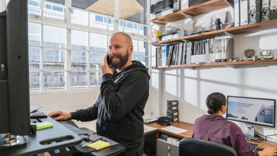 Man talking on mobile phone while working at a desktop computer, at a stand-up desk in an office.