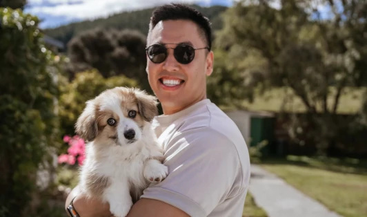 Man holds small dog in his arms while standing in a sunny garden.