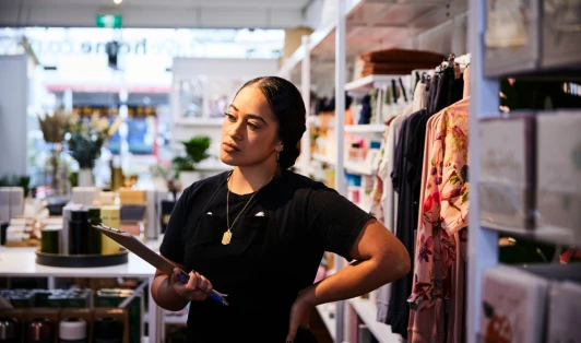 A retail worker stands in a shop, holding a clipboard.