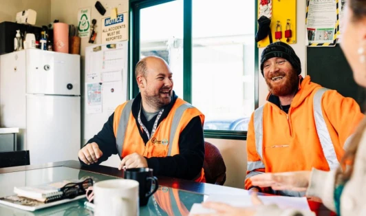 Two men in orange high-vis gear, laughing in a workplace breakroom.