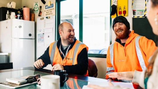 Two men in orange high-vis gear, laughing in a workplace breakroom.