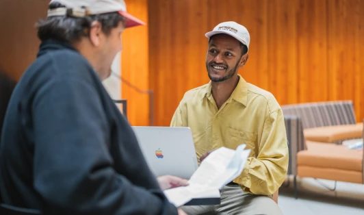 Two men chatting at a table, with a laptop and papers.
