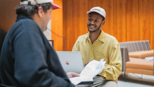 Two men chatting at a table, with a laptop and papers.