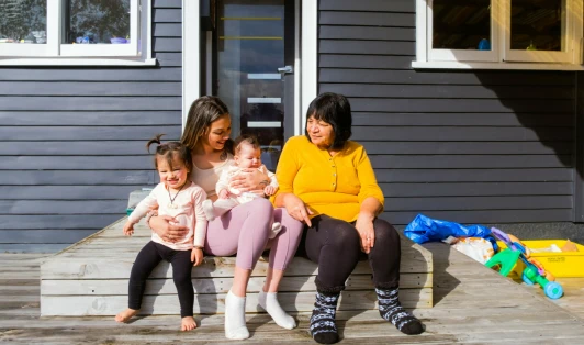 A young woman sits on a doorstep with a toddler and baby on her lap while an older woman sits next to her.