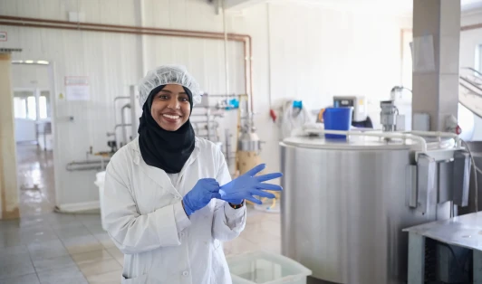Woman dressed in a white coat with gloves and hairnet stands in a food manufacturing site.