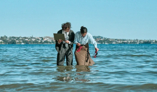 Two people in office clothes knee-deep in the sea, holding a laptop and a pair of red shoes