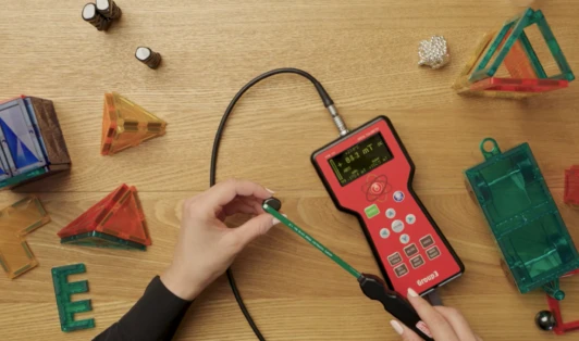 A wooden bench top covered with magnetic toys, showing a person's hands using instruments to measure quality of the toys.