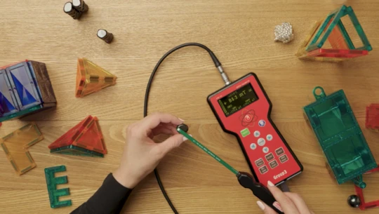 A wooden bench top covered with magnetic toys, showing a person's hands using instruments to measure quality of the toys.