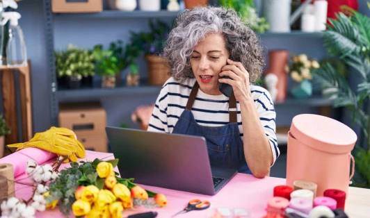 A florist speaking on a phone while looking at a laptop.
