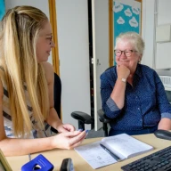An older woman smiles at a younger woman sitting at a computer desk.